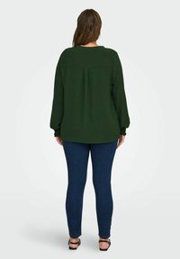 Donna in piedi di spalle con una blusa a maniche lunghe verde scuro, jeans blu scuro e sandali neri con cinturini alla caviglia.