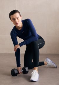Crni heksagonalni dumbbell drži osoba u mornarskoplavoj sportskoj majici dugih rukava i crnim tajicama, s bijelim detaljima i bijelim cipelama.