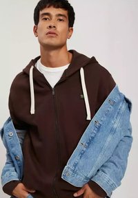 Brązowy hoodie na zamek z białymi sznurkami, noszony pod jasnoniebieską dżinsową kurtką z postarzanymi detalami i srebrnymi guzikami.