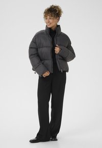 Sivi puffer kaput s visokim ovratnikom, s kondo dizajnom, zatvaranjem na zip i elastičnom donjom rubu. Nosa se preko crnog topa i hlača.