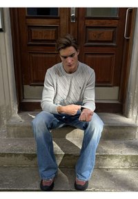 Fiatal férfi betonlépcsőn ülve, világosszürke pulóvere ujját igazítja, kék farmert és barna cipőt visel.