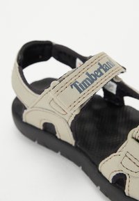 Beige lederen sandaal met zwarte accenten, verstelbare banden, getextureerde binnenzool en rubberen buitenzool, met Timberland merknaam op de band.