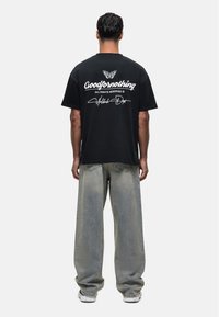 Czarna bawełniana koszulka z białym napisem i grafiką motyla na plecach, zestawiona z jasnymi, spranymi dżinsami o szerokich nogawkach i białymi trampkami.