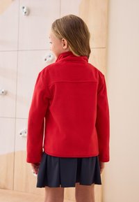 Giacca in pile rossa con collo alto, maniche lunghe e taglio dritto, indossata sopra una gonna nera plissettata. Tessuto liscio, dettagli minimali.