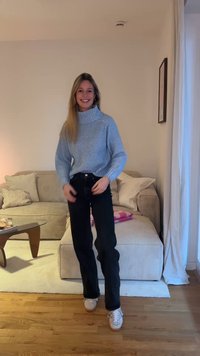 Pull à col roulé bleu clair en tricot avec manches côtelées, associé à un jean noir taille haute et des baskets blanches dans un cadre intérieur chaleureux.