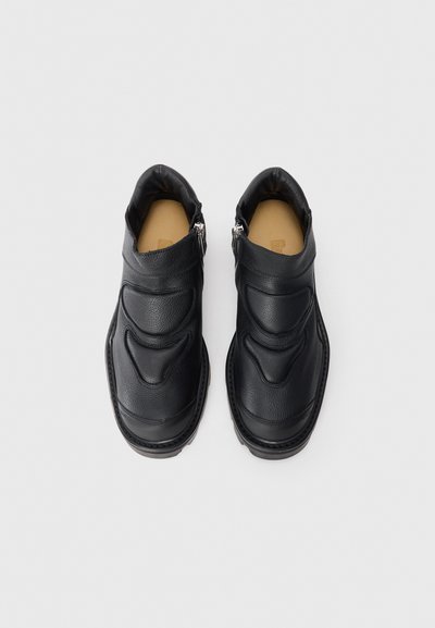 Zapatos de piel negra sin cordones con una superficie texturizada, punta redondeada y cierre de cremallera; cuentan con un cuello acolchado y una suela marrón contrastante.