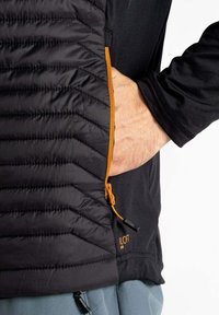Schwarze isolierte Jacke mit gestepptem Material und orangefarbigem Reißverschluss. Verfügt über eine Seitentasche mit Reißverschlussdetails und kontrastierendem Stoff darunter.