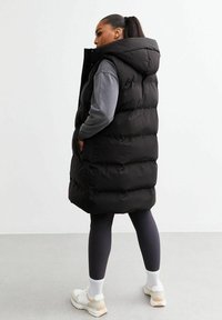 Gilet imbottito nero con cappuccio, caratterizzato da un design trapuntato e tasche laterali. Indossato sopra un top grigio a maniche lunghe e leggings neri. Sneakers bianche.