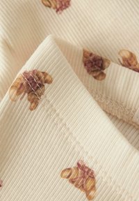 Geribbelde katoenen stof in roomkleur met verspreide bloemenpatronen in gedempt bruin en bordeauxrood. Kenmerkt contrasterende stiksels langs de randen.