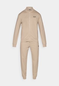 Bēša sporta tērps no mīksta auduma, kurā ir rāvējslēdzēja jaka ar augstu apkakli un bikses ar manšetēm. Uz krūtīm ir uzdrukāts logotips.