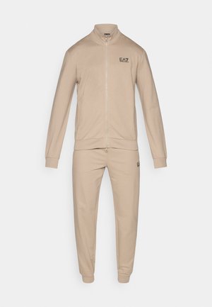 Beige tracksuit made of soft fabric, featuring a zip-up jacket with a high collar and pants with cuffs. Includes a logo on the chest.
