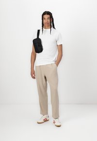 Camiseta branca, calças largas bege, mala de ombro preta com fecho, sapatos cremosos com riscas vermelhas, usada por um indivíduo em pé.