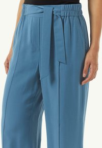 Jasnoniebieskie spodnie o gładkiej fakturze, z elastycznym pasem i wstążką z przodu. Zawierają boczne kieszenie.