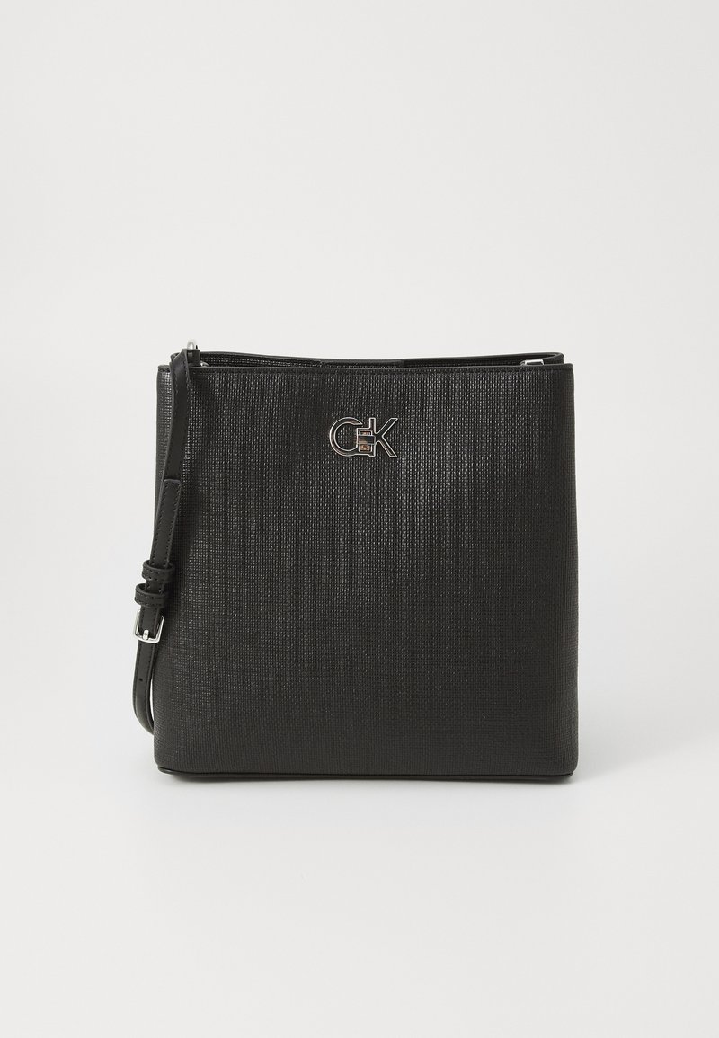 Svart crossover-veske med teksturert ytre, flat form, glidelås lukking, og sølvfarget logo aksent på forsiden. Justerbar skulderrem inkludert.