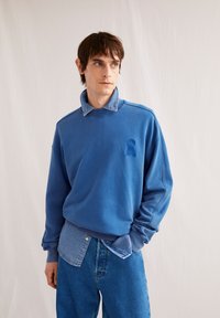 Blaues Sweatshirt mit strukturiertem Kragen, versehen mit einem blauen gestickten Detail. Kombiniert mit hellblauer Jeans, was einen geschichteten Look kreiert.