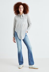 Camisa de botones a rayas gris claro con un bolsillo en el pecho y bordado de hojas de palma, combinada con jeans acampanados azul desgastado y zapatillas ligeras.