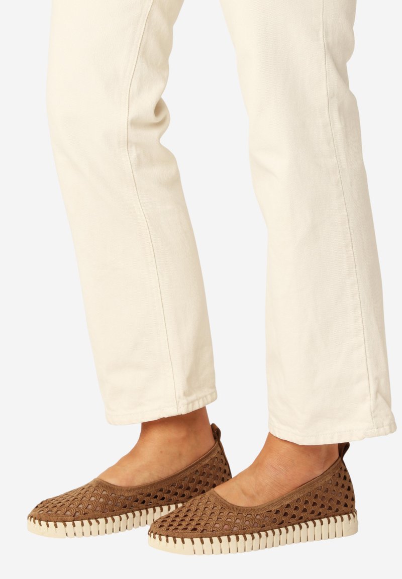 Voeten met bruine gevlochten instapschoenen met witte zolen, gecombineerd met crèmekleurige rechte broekspijpen.
