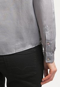 Camicia grigia con texture e polsini a bottoni, vista dal retro; abbinata a pantaloni neri con tasca posteriore e dettagli di cucitura.