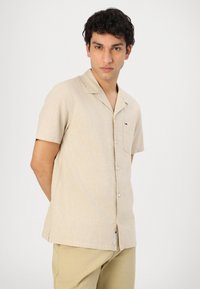 Kortermet, beige skjorte laget av lett stoff med en subtil tekstur, med knappestolpe og en liten logo på brystlommen.