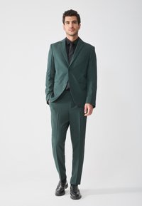 Dunkelgrüner Anzug mit einreihigem Blazer und geraden Hosen, kombiniert mit einem schwarzen Hemd und Schuhen. Glatte Stoffoberfläche.