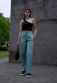 Top cropped preto de uma só manga, calças com faixa elástica em teal claro e sapatos pretos. Modelo segura uma bolsa texturizada preta, em pé contra uma parede cinza.