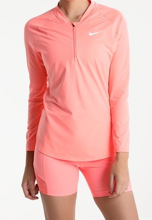 Frau trägt ein leuchtend korallenrotes, langärmliges Sportoberteil mit Viertelreißverschluss und passende enganliegende Shorts vor weißem Hintergrund.