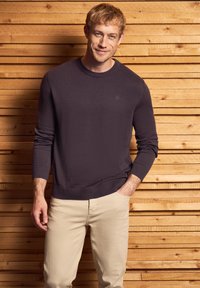 Dunkelvioletter Rundhals-Pullover aus Strickstoff, mit gerippten Bündchen und Saum, kombiniert mit hellbeigen Hosen, vor einem Holz-Hintergrund präsentiert.