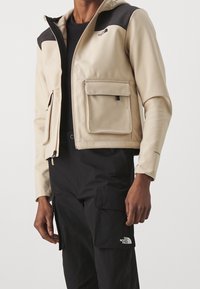 Beige bunda s černými ramenními akcenty, dvěma předními kapsami, zipovým zapínáním a kapucí. Nositá přes černé cargo kalhoty.