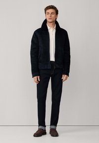 Navy nubuck jacket met hoge kraag, knoopsluiting aan de voorkant en zijzakken. Gepaard met donkere spijkerbroeken en bruin instap schoenen.