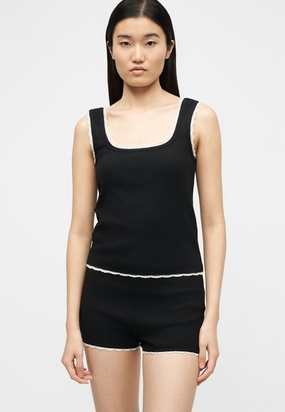 Femme portant un débardeur noir à côtes et un short assorti avec un bord en dentelle blanche, se tenant devant un fond clair uni.