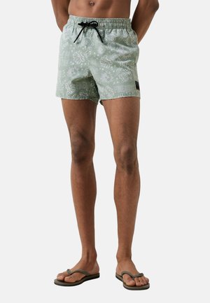 Man draagt lichtgroene zwemshorts met witte tropische patronen en een zwart trekkoord, gecombineerd met donkergroene teenslippers, staand tegen een witte achtergrond.