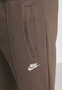 Hnědé Nike tepláky s měkkou texturou, které mají boční kapsu a bílé logo značky blízko kapsy.