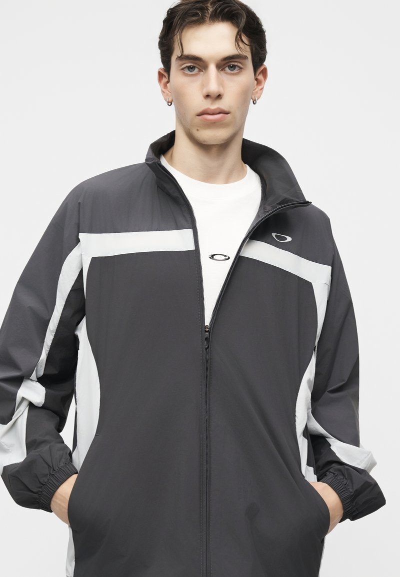 Joven con una chaqueta con cremallera en blanco y negro y camisa blanca, de pie con las manos en los bolsillos frente a un fondo liso.