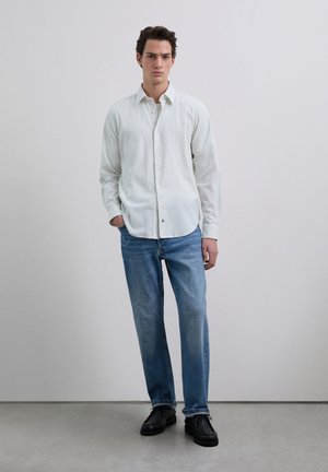 Camisa abotonada con rayas verticales en colores claros; vaqueros de corte holgado; zapatos negros con cordones; diseño minimalista sobre un fondo neutro.