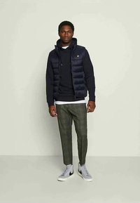 Bărbat în picioare, îmbrăcat cu o vestă căptușită de culoare bleumarin peste un hanorac negru, pantaloni cu carouri verzi și adidași gri cu accente negre.