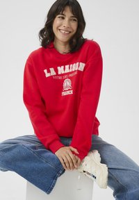 Rotes Sweatshirt mit weißer Aufschrift: "LA MAISON VOTRE LIEU PRÉFÉRÉ FRANCE", kombiniert mit hellblauen, weit geschnittenen Jeans und weißen Turnschuhen.