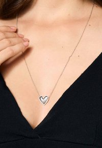 Srebrny naszyjnik w kształcie serca z wypolerowanym wykończeniem, zawieszony na cienkim łańcuszku. Noszony na tle czarnej tkaniny.