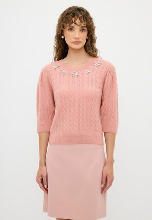 Pull en tricot rose clair avec des manches courtes, présentant un motif texturé et des embellissements floraux décoratifs au niveau de l'encolure.