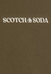 Oliven grønt stof med ribbet tekstur, der har det hvide broderede mærkenavn "SCOTCH & SODA" i en fed, moderne skrifttype.