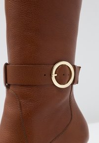 Botte haute jusqu'au genou en cuir marron avec une large bride et une boucle circulaire dorée, surface texturée. Couture lisse le long des bords.