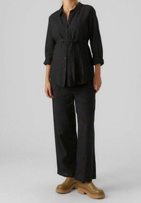 Czarna lniana koszula z guzikami i podwiniętymi rękawami, z paskiem w talii, w połączeniu z dopasowanymi czarnymi spodniami o szerokich nogawkach i masywnym, beżowym obuwiem.