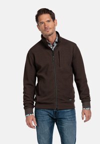 Bruine zip-up jack met een gestructureerde stof, staande kraag en zijzakken. Gecombineerd met blauwe jeans en een bloemenoverhemd eronder.