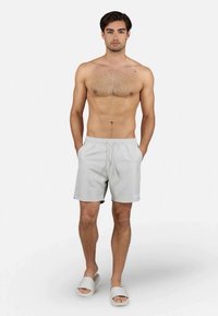 Joven de pie, sin camisa, vistiendo un bañador gris claro y sandalias a juego contra un fondo blanco liso.