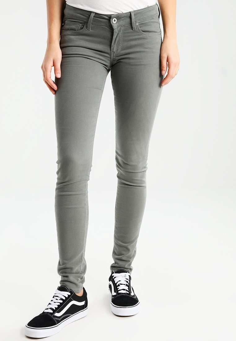 Femme portant un pantalon gris slim fit et des baskets noir et blanc, debout avec les mains détendues le long du corps sur un fond blanc.