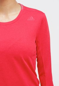 Czerwona sportowa koszulka z długim rękawem o teksturowanym wykończeniu, z subtelnym logo Adidas na lewym ramieniu i okrągłym dekoltem.