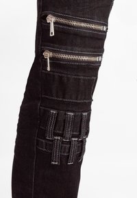 Pantaloni in denim neri con più tasche con zip e un pannello testurizzato con dettagli cuciti sulla coscia. Dettagli in metallo argentato.