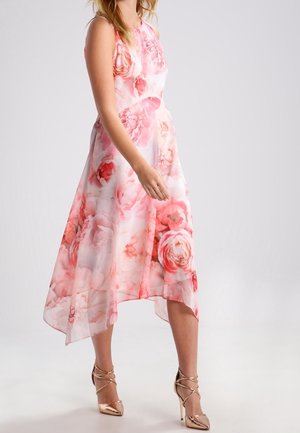 Vrouw draagt een mouwloze roze-witte gebloemde jurk met asymmetrische zoom en gouden hoge hakken met bandjes.