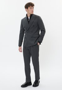Šedý dvouřadý oblek s černými knoflíky, na míru šitý střih, spárován s černým zipovým topem a lesklými černými botami. Jednoduché pozadí.