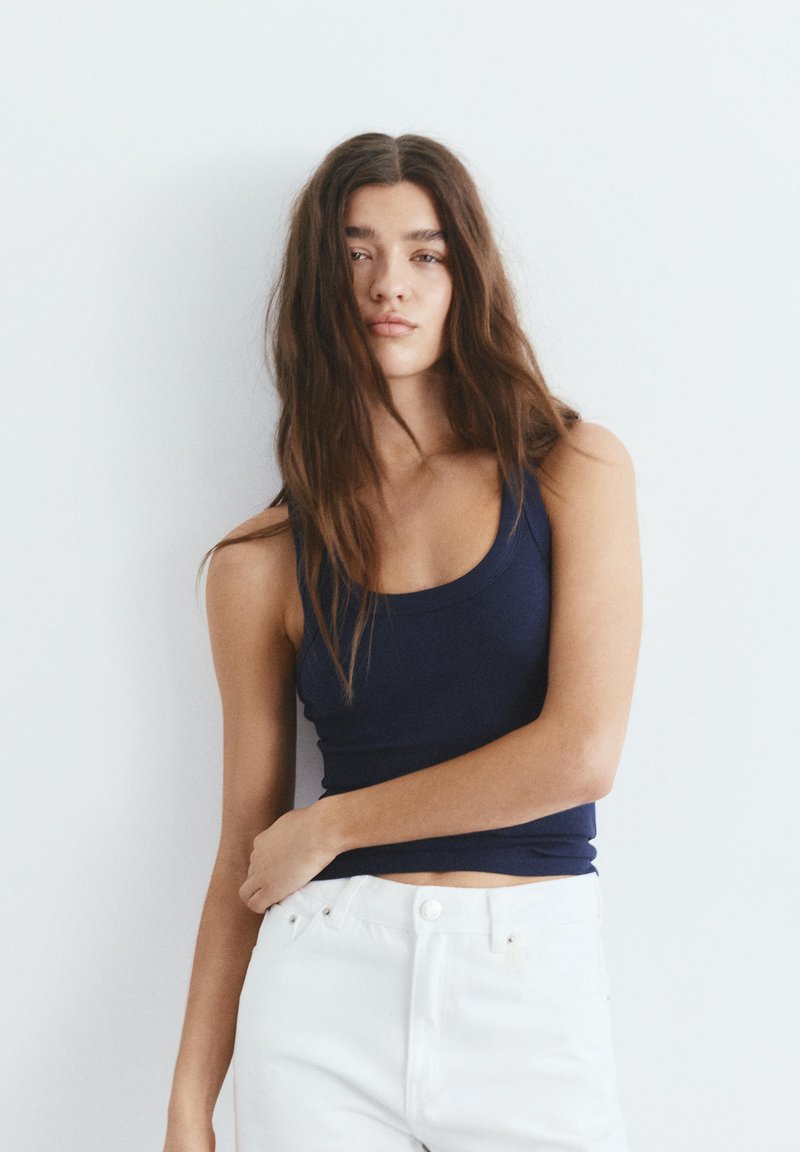 Jeune femme aux longs cheveux bruns portant un débardeur bleu marine et un pantalon blanc, debout devant un fond uni gris clair.