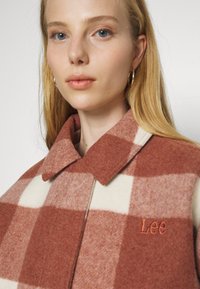 Kostkovaný vlněný kabát v rezavé a krémové barvě, vybavený límcem, vyšívaným logem "Lee" a texturovanou tkaninou. Design obsahuje velké čtverce.